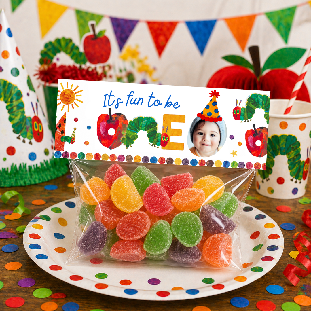 Hungry Caterpillar themed candy favor bag with colorful gumdrops inside a clear cellophane pouch, topped with a personalized “It’s fun to be ONE” printable bag topper, displayed on a festive party table with rainbow bunting, confetti, and themed party cups.
