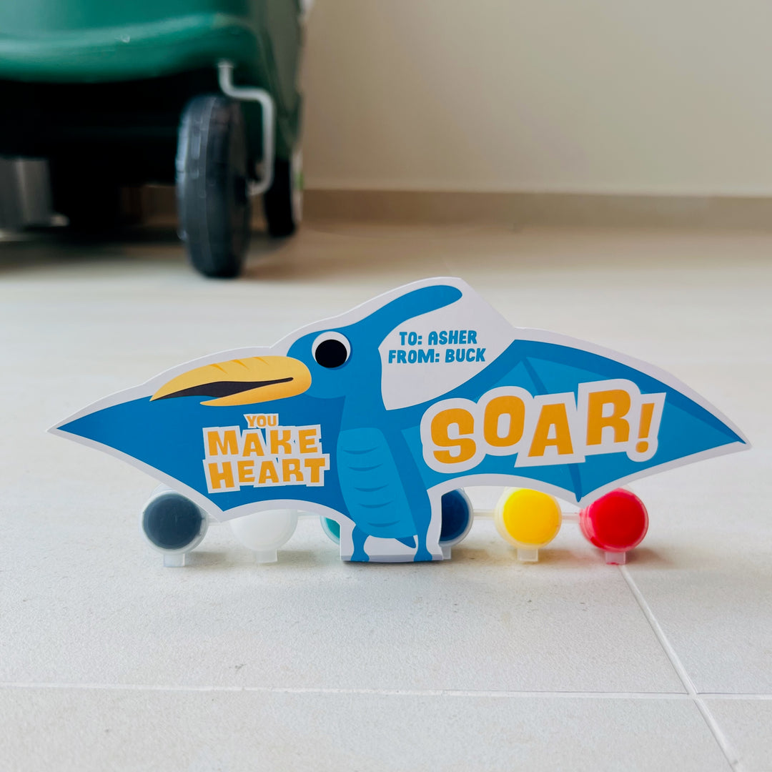 Printable flying dinosaur gift holder in blue pterodactyl design with "You Make My Heart Soar!" and paint tube inserts for valentines classroom gift exchanges