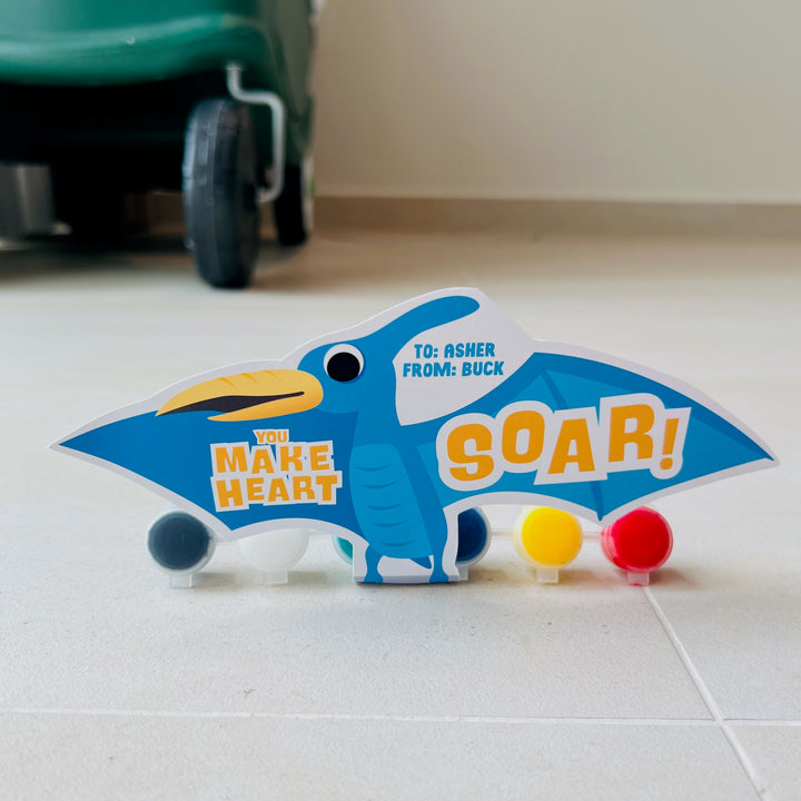 Printable flying dinosaur gift holder in blue pterodactyl design with "You Make My Heart Soar!" and paint tube inserts for valentines classroom gift exchanges