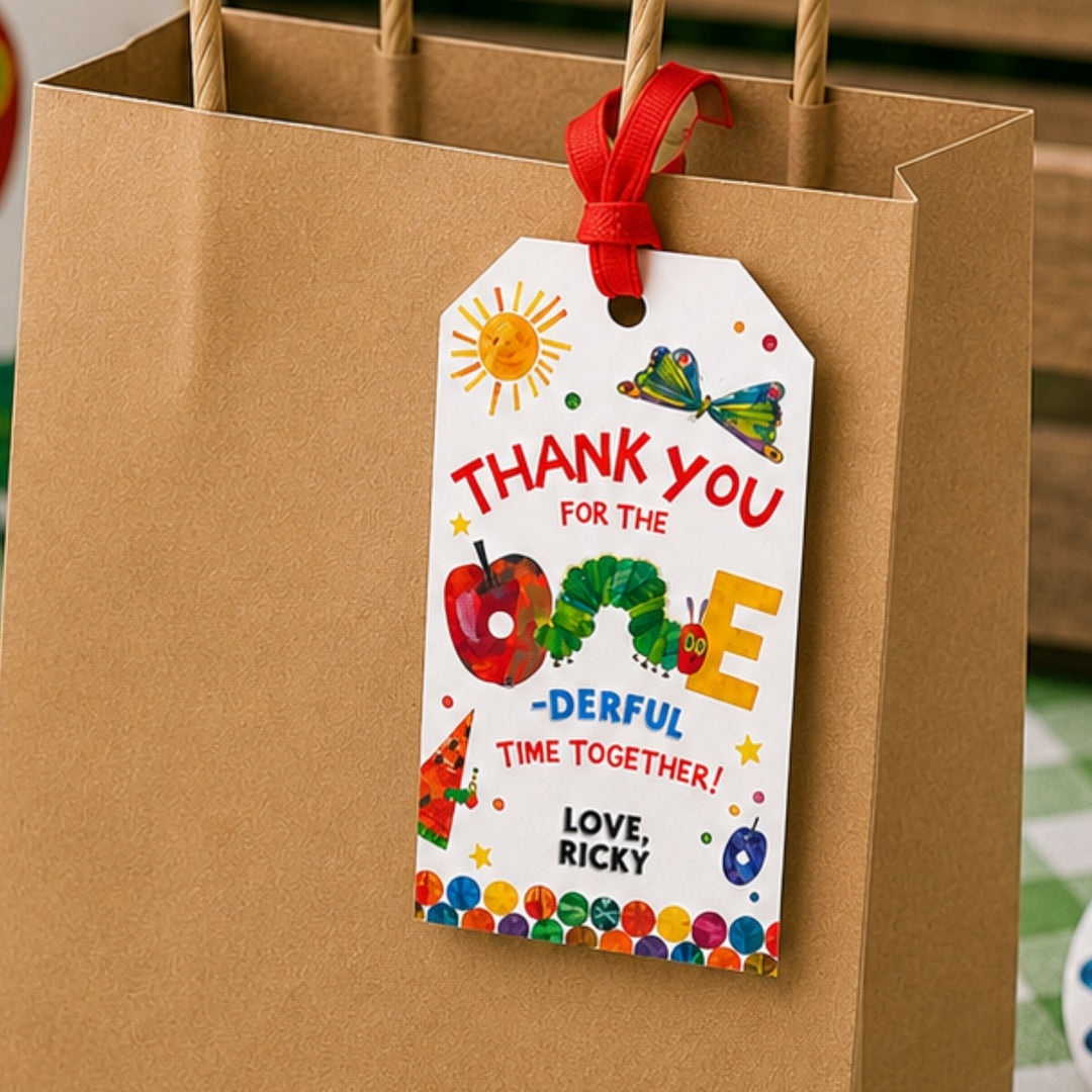 Editable Very Hungry Caterpillar gift tag printable attached to a kraft paper favor bag with red ribbon, featuring Eric Carle's very hungry caterpillar, butterfly illustrations, and “ONE-derful” birthday wording for first birthday party favors and thank-you bags.